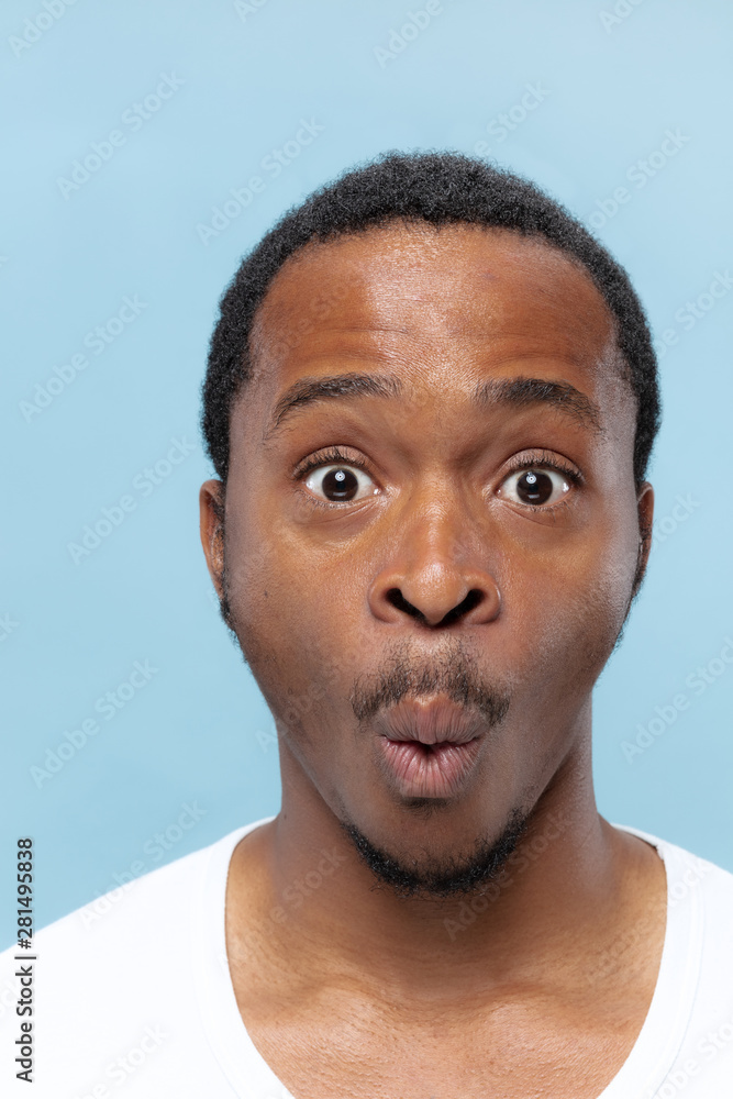 Close up portrait of young african-american man in white shirt on blue background. Human emotions, facial expression, ad, sales concept. Looking at camera, looks shocked, astonished, wondered. Close up portrait of young african-american man in white shirt on blue background. Human emotions, facial expression, ad, sales concept. Looking at camera, looks shocked, astonished, wondered.