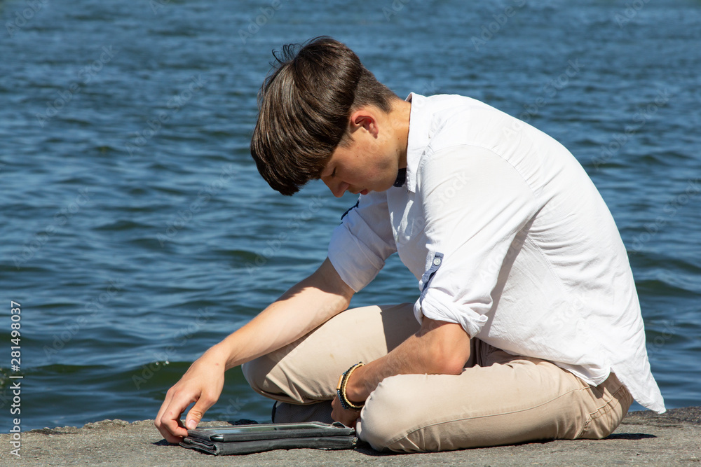 Portrait of a teenager, man, in a white shirt against a blue sky and a large lake. A high school student sits by the lake with a tablet and reads. The young man looks tired.