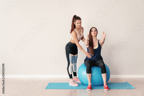 Young sporty woman with fitball doing exercises under supervision of her personal trainer near light wall