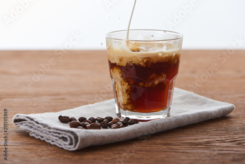 milk pouring into ice coffee in glass on napkin with coffee grains on wooden table isolated on white