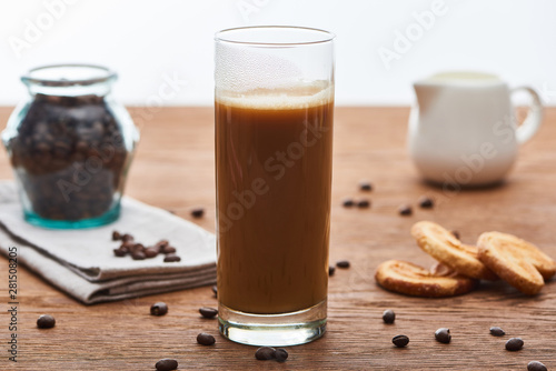 selective focus of ice coffee with milk in glass near milk jug, cookies and coffee grains on wooden table isolated on white