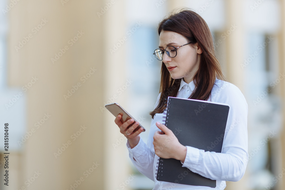 Portrait young business woman wearing white shirt using smartphone out doors. Female reading sms message in working process. Portrait young business woman wearing white shirt using smartphone out doors. Female reading sms message in working process.