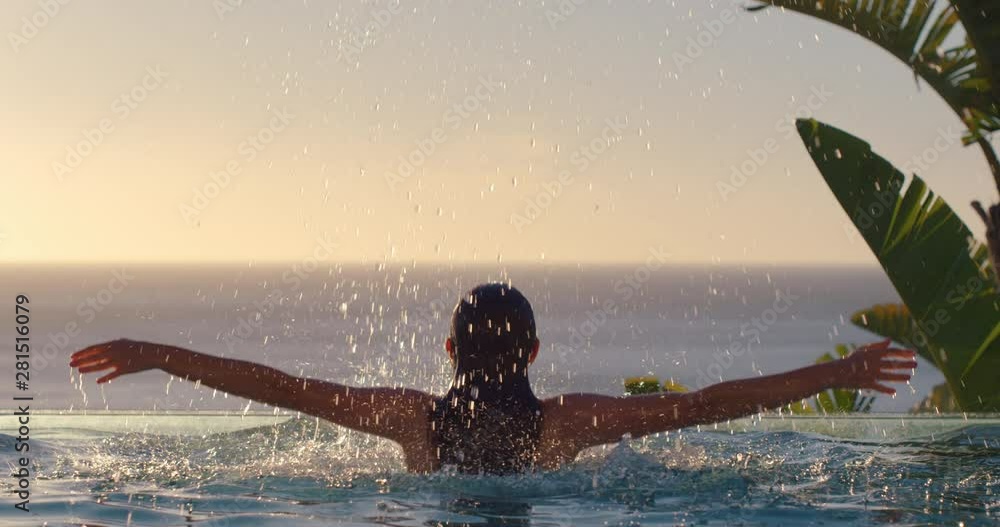 happy woman jumping out of swimming pool water with arms raised enjoying summer vacation in mediterranean hotel resort with beautiful view of ocean at sunset travel freedom 4k
