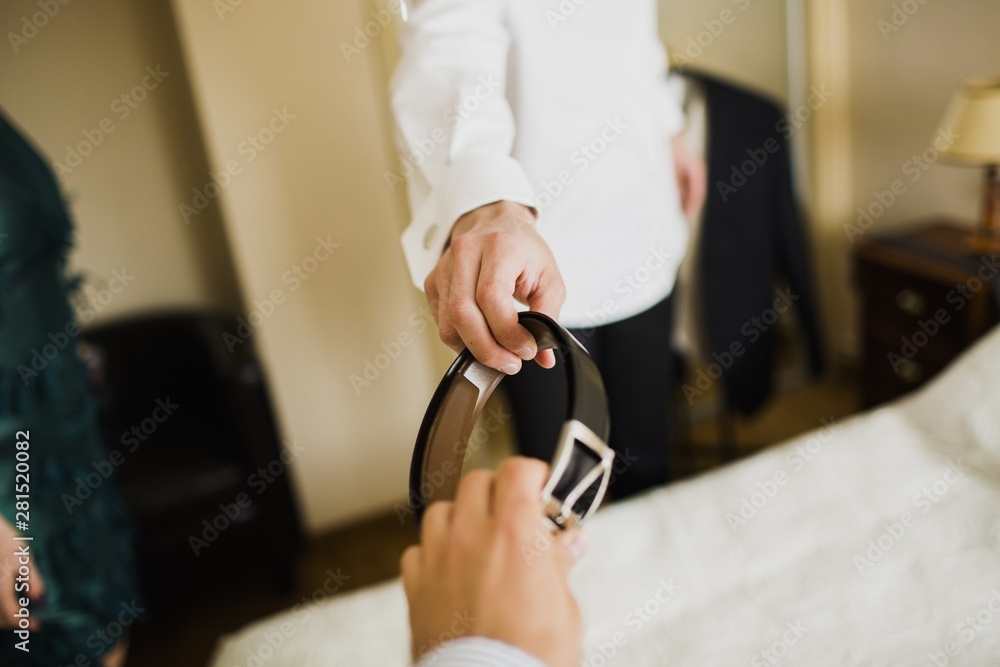 A man wears his wedding suit, while someone passes his belt with his hand.
