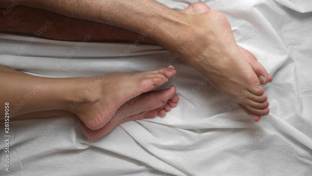 Top view. Female and male legs in bed on a white sheet caress each other. Romance at home. Close-up. Top view. Female and male legs in bed on a white sheet caress each other. Romance at home. Close-up.