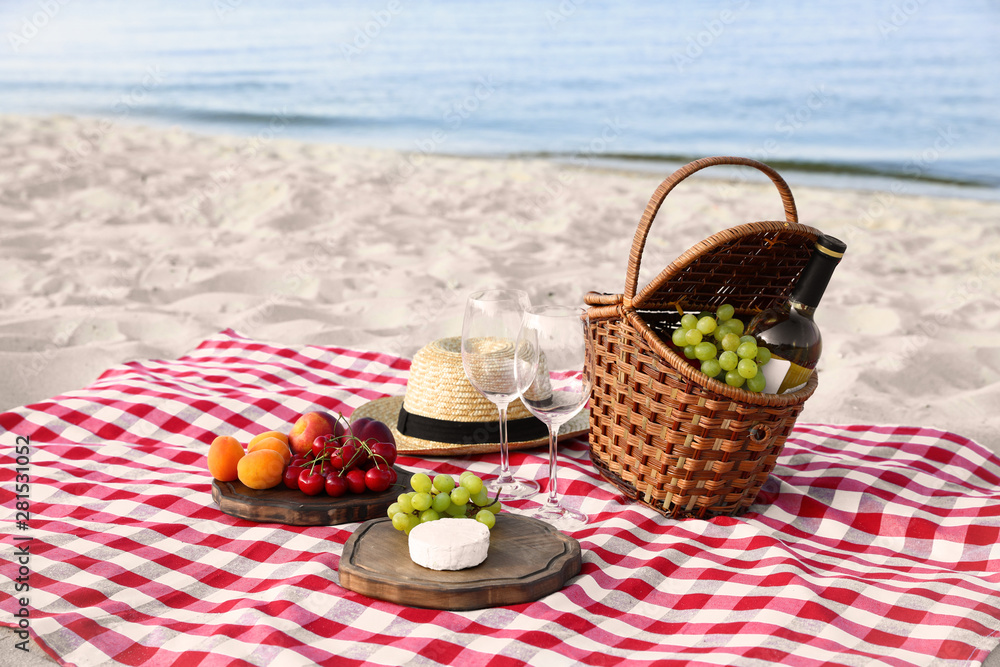 Checkered blanket with picnic basket and products on sunny beach Checkered blanket with picnic basket and products on sunny beach