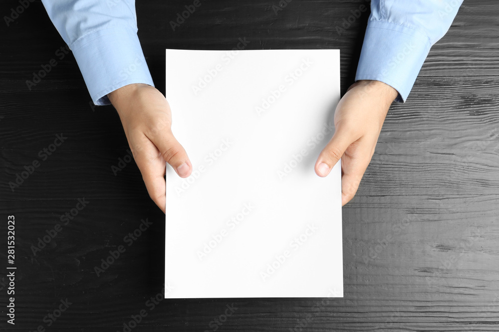 Man holding blank paper sheet for brochure at black wooden table, top view. Mock up Man holding blank paper sheet for brochure at black wooden table, top view. Mock up