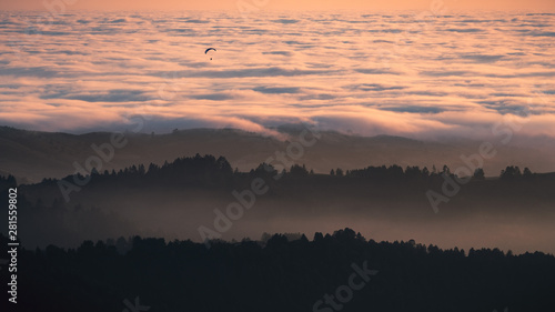 Sunset view of layered hills and valleys covered by a sea of clouds in Santa Cruz mountains; paraglider visible above the clouds; San Francisco bay area, California