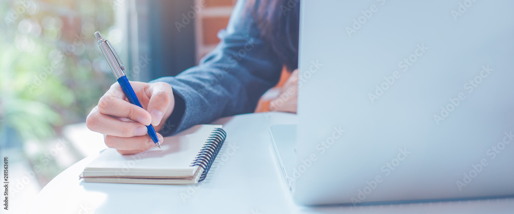 Woman hand is writing on a notepad with a pen and have a laptop computer on the desk in the office.Web banner.