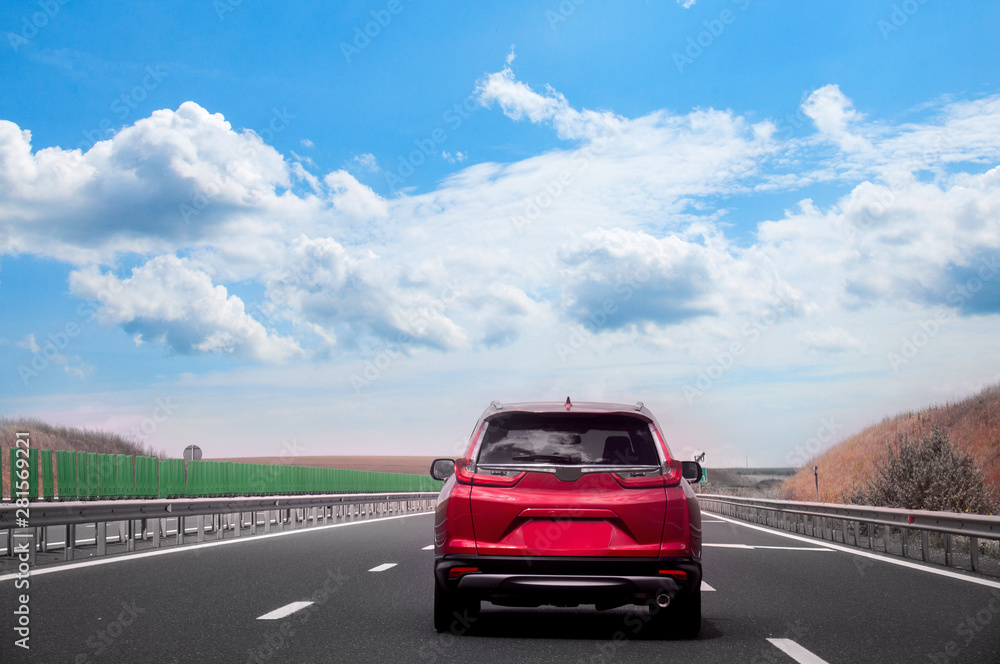 Fototapeta premium The red car is racing along a straight road against the background of a golden field and a blue sky. Travelling by car.