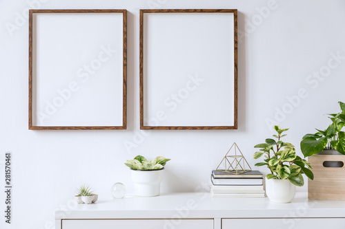Elegant home interior with two brown wooden mock up photo frames above the white shelf with books, plants, gold pyramid, wooden box and home accessories. Stylish concept of white room decor. 