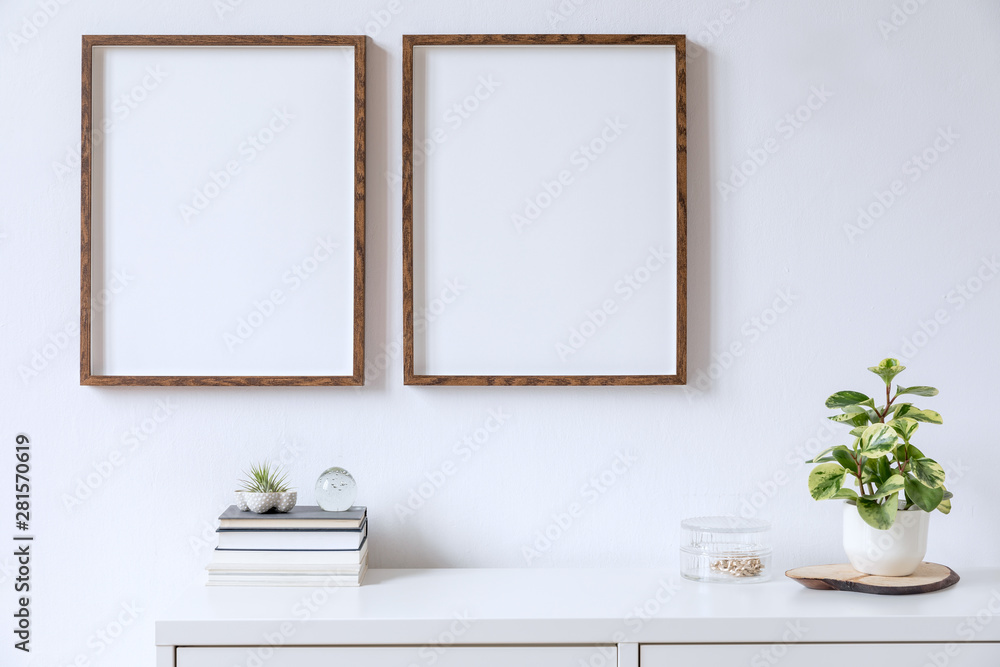 Fototapeta Minimalistic home decor of interior with two brown wooden mock up photo frames above the white shelf with books, beautiful plant in stylish pot and home accessories. White wall. Concept of mockup.