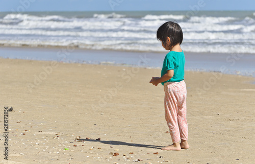 Asian little girl with short hair wearing green shirt standing alone on the beach nearby the sea staring on the sandy ground in the afternoon