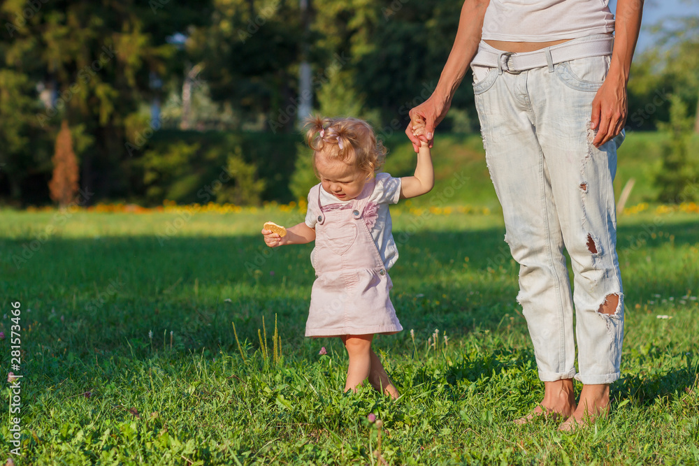 Little cute girl does her first steps with mother support, green grass, summer time, blurred background Little cute girl does her first steps with mother support, green grass, summer time, blurred background