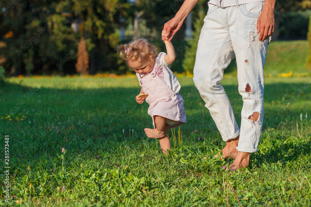 Little cute girl does her first steps with mother support, green grass, summer time, blurred background Little cute girl does her first steps with mother support, green grass, summer time, blurred background