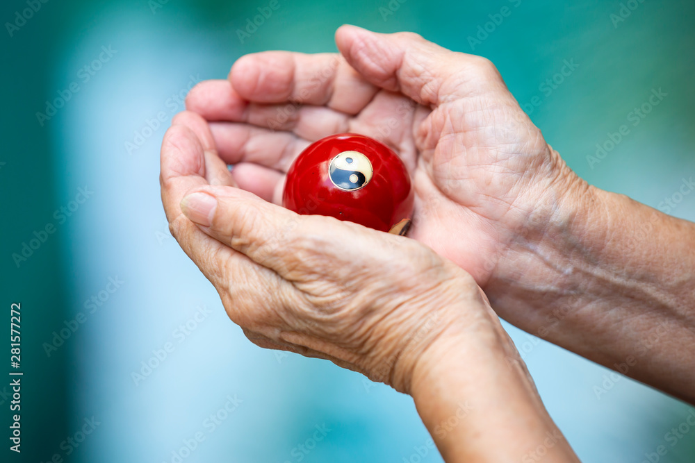 Senior woman's hands holding and praying a black and white Yin Yang religious symbol on red ball in Taoism in bokeh blue swimming pool background, Asian Body language feeling, Religion concept Senior woman's hands holding and praying a black and white Yin Yang religious symbol on red ball in Taoism in bokeh blue swimming pool background, Asian Body language feeling, Religion concept