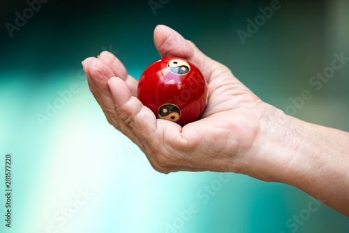 Senior woman's hand holding and praying a black and white Yin Yang religious symbol on red ball in Taoism in bokeh blue swimming pool background, Asian Body language feeling, Religion concept