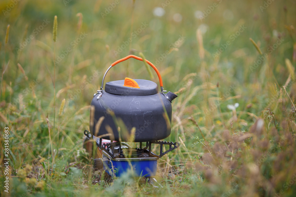 A small grey camping teapot stands on the grass. Use in the campaign of special camping utensils. A small grey camping teapot stands on the grass. Use in the campaign of special camping utensils.