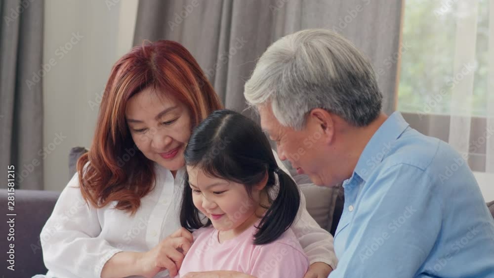 Asian grandparents and granddaughter using tablet at home. Senior Chinese, grandpa and grandma happy spend family relax with young girl check social media, lying on sofa in living room. Slow motion. Asian grandparents and granddaughter using tablet at home. Senior Chinese, grandpa and grandma happy spend family relax with young girl check social media, lying on sofa in living room. Slow motion.