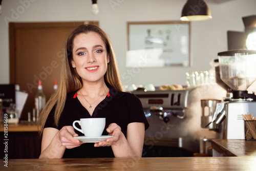 Beautiful female barista is working in coffee shop. Attractive woman is standing behind the bar counter  making coffee and welcomes customers.