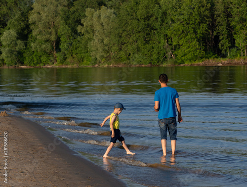 Father and son walk on the river. A man stands in the water and looks at the waves, his son plays with the waves. Sunny day. Back view. On the other side of the river is a forest.
