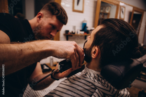 Getting perfect shape. Close-up side view of young stylish bearded man getting beard haircut by bearded, muscular hairdresser or barber at barbershop