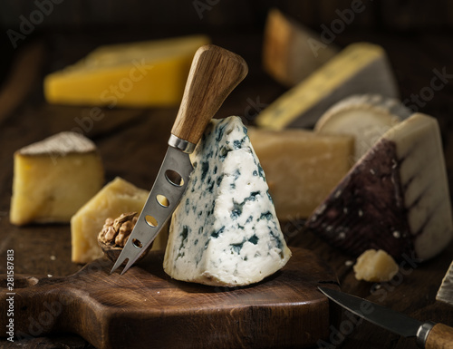 Segment of blue mould cheese and cheese knife on wooden board. Different cheeses at the background.