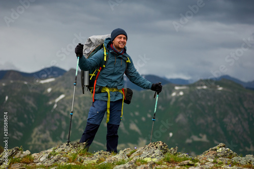Smiling hiker man stands on the cliff against green mountain range. Man has a backpack and trekking poles. Mountain trekking. Copy space © kravtzov