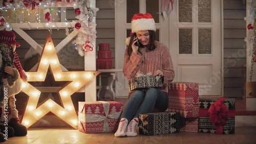 Young attractive girl in santa cap and sweater wrapping christmas gifts, presents sitting at house porch and speaking mobile phone. Christmas concept. Full length sshot with copy space photo