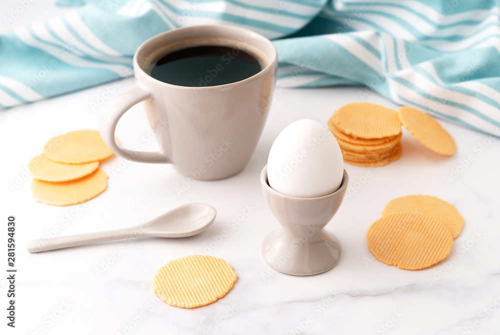 Boiled egg in ceramic egg cup, cup of coffee and thin crispy corn chips on background of turquoise kitchen towel. Breakfast concept. Boiled egg in ceramic egg cup, cup of coffee and thin crispy corn chips on background of turquoise kitchen towel. Breakfast concept.