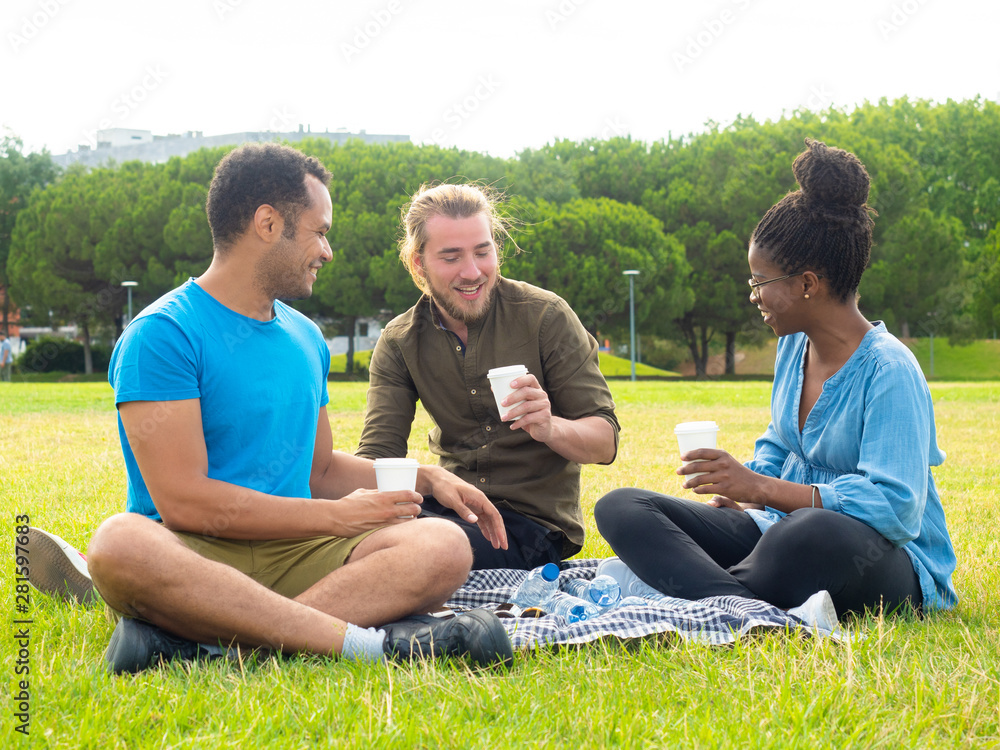 Cheerful friends holding paper cups in park. Smiling young multiethnic friends sitting on plaid and talking in park. Weekend concept