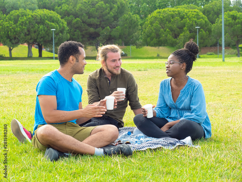 Multiethnic friends holding paper cups and talking in park. Three happy male and female friends sitting on green lawn and talking outdoor. Friendship concept