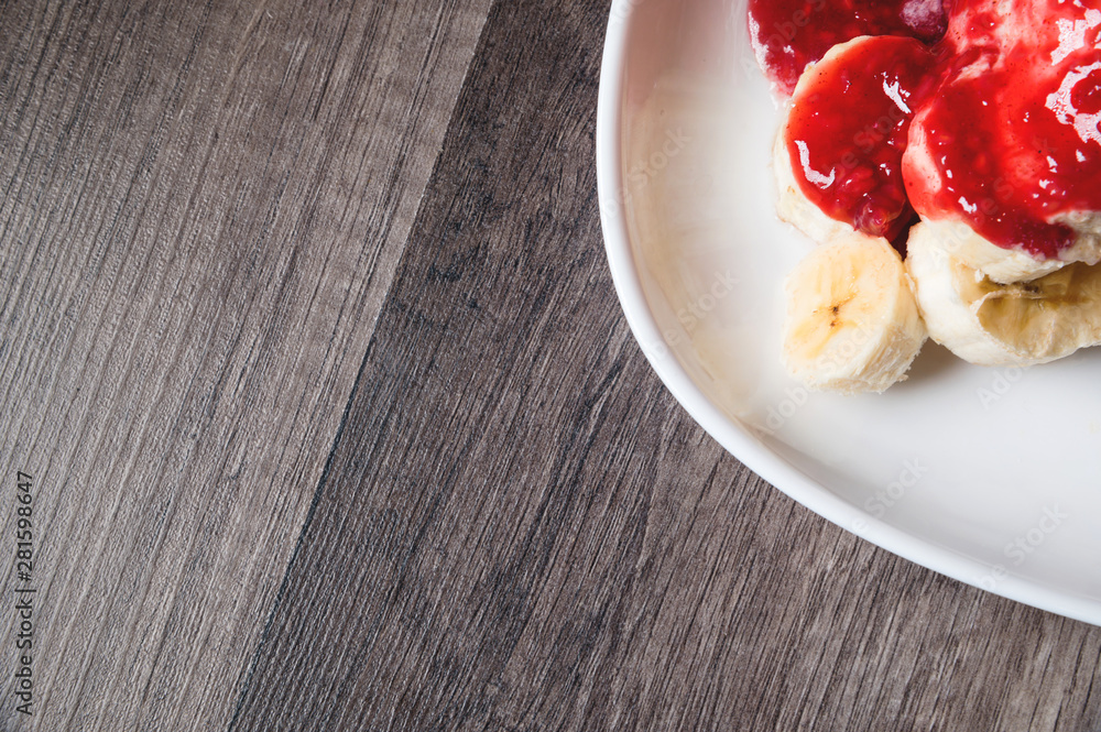 Closeup Vanilla ice cream poured with strawberry or raspberry jam with sliced banana slices on a white plate on a dark wooden table. You can do this at home on vacation Closeup Vanilla ice cream poured with strawberry or raspberry jam with sliced banana slices on a white plate on a dark wooden table. You can do this at home on vacation