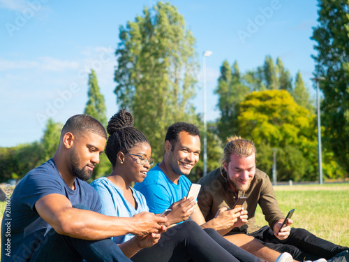 Happy friends using smartphones in park. Smiling young multiethnic friends sitting on green grass and using mobile phones. Technology concept