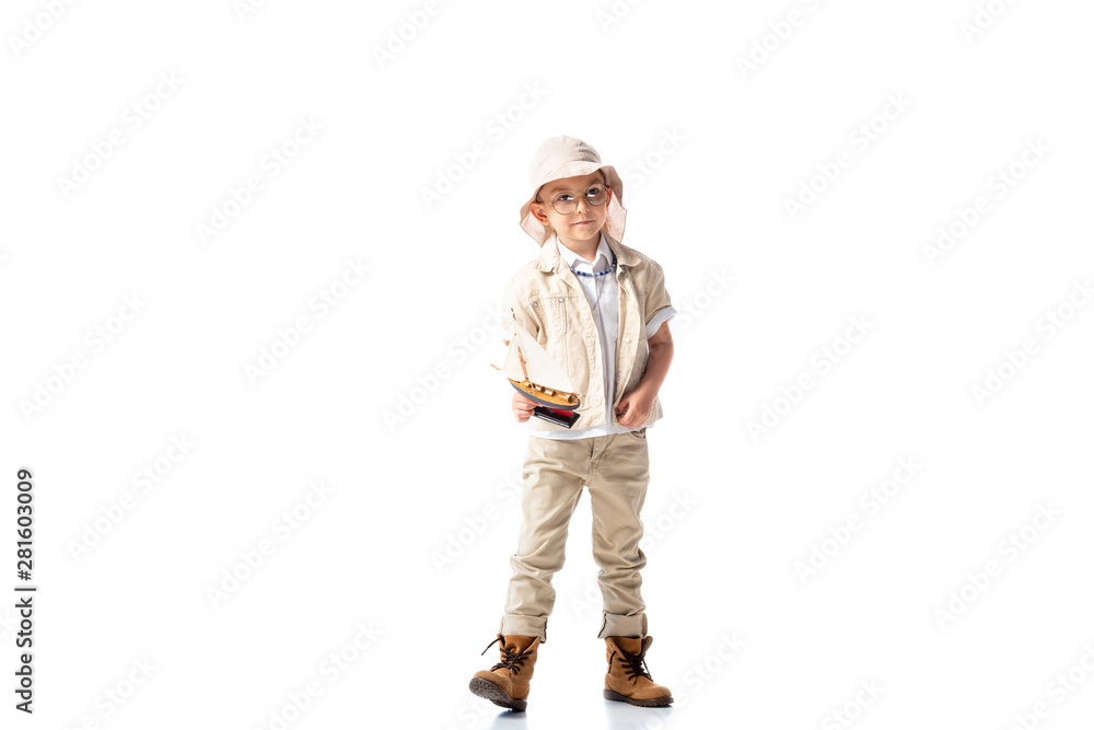 full length view of explorer child in glasses and hat holding toy ship on white