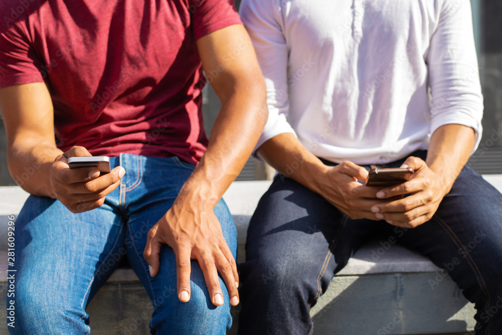 Closeup of two men in casual using smartphones outside. Cropped portrait of two male friends in casual sitting on parapet outdoors and holding mobile phones. Communication concept Closeup of two men in casual using smartphones outside. Cropped portrait of two male friends in casual sitting on parapet outdoors and holding mobile phones. Communication concept