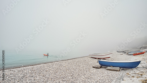 Blue and red fishing boats on a rocky pebble beach in the early morning with foggy mist on background