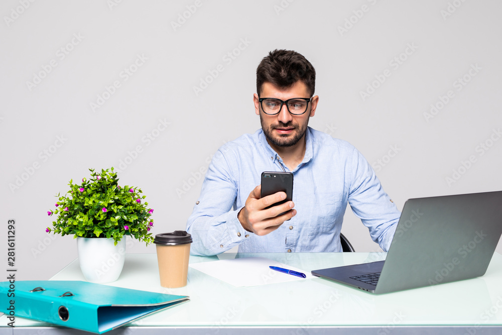 Young bearded businessman in shirt sits in office at table and uses smartphone and drinks coffee on desk with laptop. Young bearded businessman in shirt sits in office at table and uses smartphone and drinks coffee on desk with laptop.