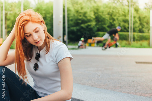 Attractive redhead smiling girl in casual clothes sitting on street in city, active people on background