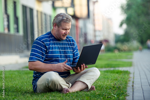 Happy man looking for online content on laptop sitting on lawn on summer day in city