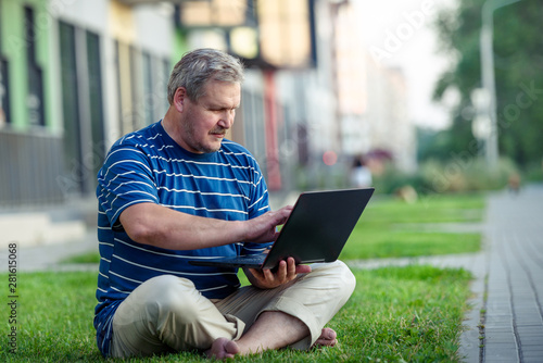 Happy man looking for online content on laptop sitting on lawn on summer day in city