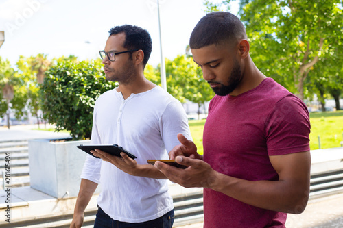 Serious mix raced guys with digital devices waiting someone. Two men standing outside, using smartphone and tablet and looking away. Communication concept
