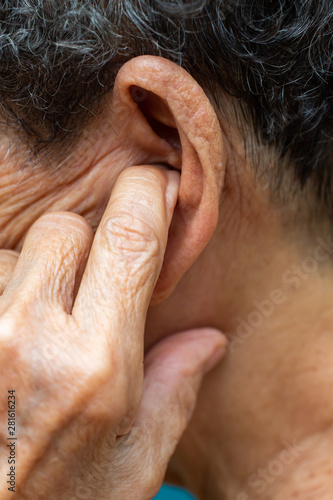 Senior woman putting index finger into her left ear, Grey curly hairs, Swimming pool background, Close up & Macro shot, Selective focus, Asian Body part, Healthcare concept, Symptom of hearing loss
