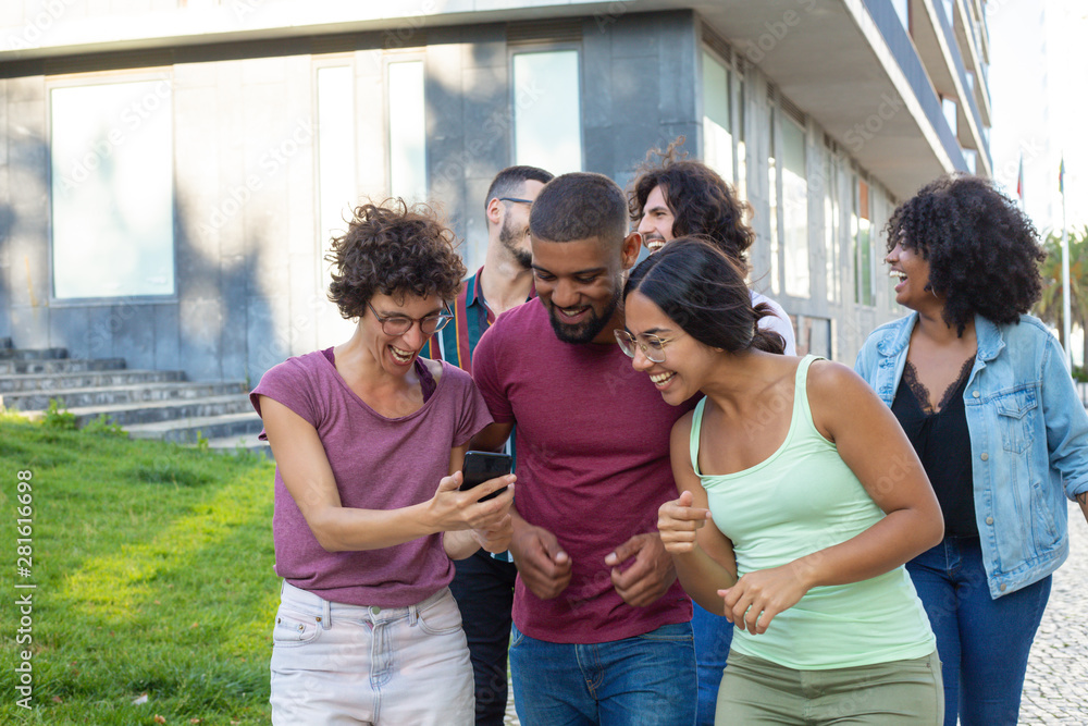 Obraz premium Joyful woman sharing good news with two happy friends and showing smartphone screen. Three laughing people walking behind them outdoors. Good news concept