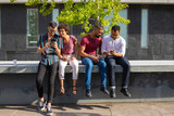 People sitting on parapet outside and watching content on phones together. Men and women using smartphone outdoors. Wireless connection concept