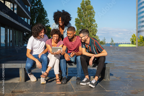 Joyful friends watching video on tablet outdoors. Interracial group of men and women sitting on bench outside, looking at tablet screen and having fun. Leisure concept