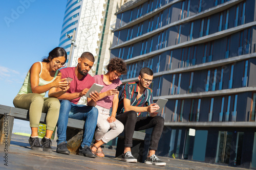 Happy young men and women using gadgets, reading on screen and watching content. People sitting on bench outdoors and using smartphones and tablets. Digital gadgets concept