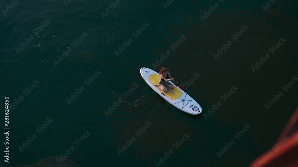 aerial view of woman sitting on a SUP paddle board and paddling rowing on a smooth calm river. evening on the river. water activities and entertainment, healthy living. slow motion aerial view of woman sitting on a SUP paddle board and paddling rowing on a smooth calm river. evening on the river. water activities and entertainment, healthy living. slow motion
