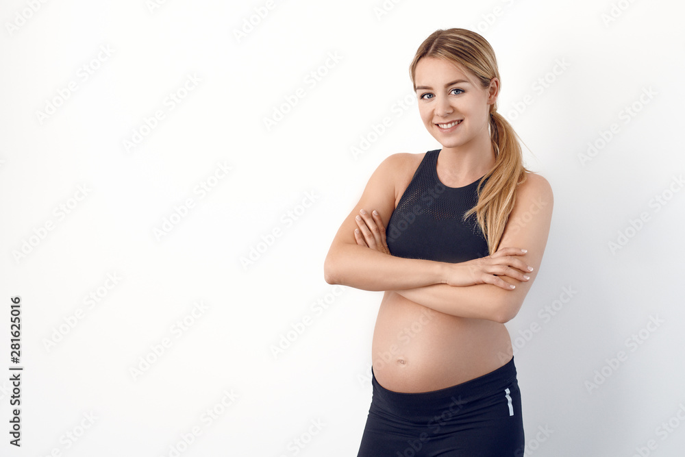 Happy healthy young pregnant woman in black sportswear standing with hands on hips looking at the camera with a beaming smile Happy healthy young pregnant woman in black sportswear standing with hands on hips looking at the camera with a beaming smile