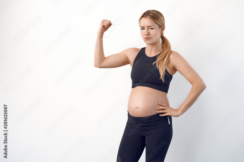 Fun loving young pregnant woman with bare baby belly standing flexing her arm to show her muscles with a wry smile over a white studio background Fun loving young pregnant woman with bare baby belly standing flexing her arm to show her muscles with a wry smile over a white studio background